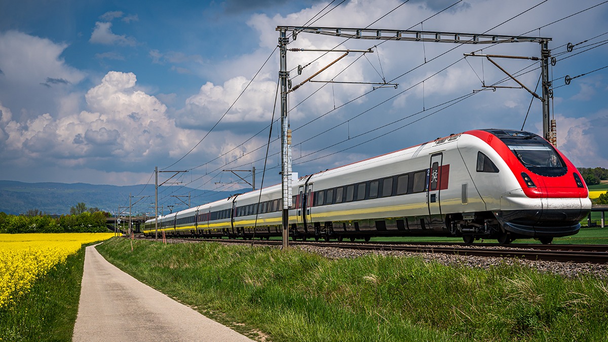 Train running through rapeseed field in spring. One red electric train, railroad with blue sky, yellow flowers and green field. Chavornay, Switzerland. Transport.